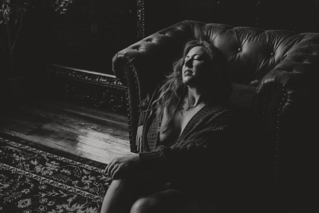 Moody black and white boudoir image of a woman relaxing by a vintage chair, taken at Elevation Boudoir in Tuscaloosa, AL.