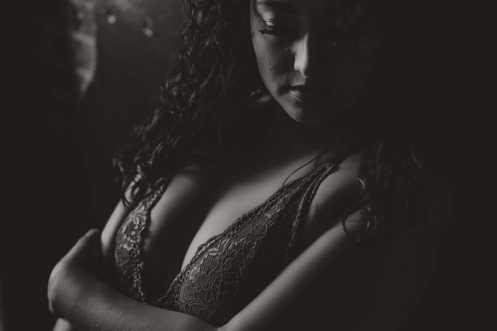 Black and white boudoir image of a woman in lace lingerie with arms wrapped around her body, quietly powerful and reflective.