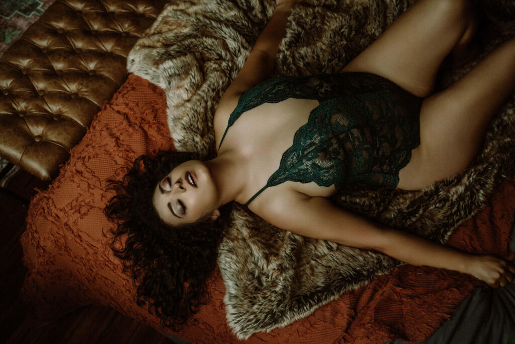 Full-body portrait of a woman in green lace bodysuit lying on a rust-colored blanket and faux fur, eyes closed, symbolizing softness and personal renewal.