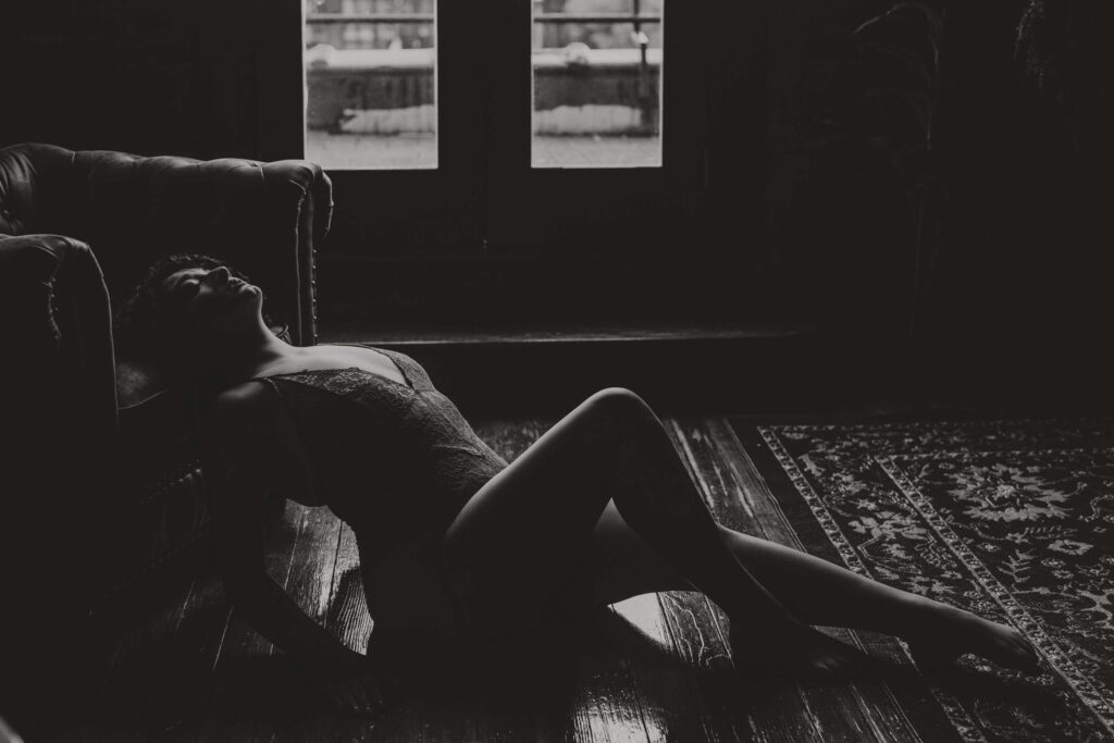 Moody black and white silhouette of a woman in lingerie seated on the floor near double doors, light framing her form in a moment of solitude and strength.
