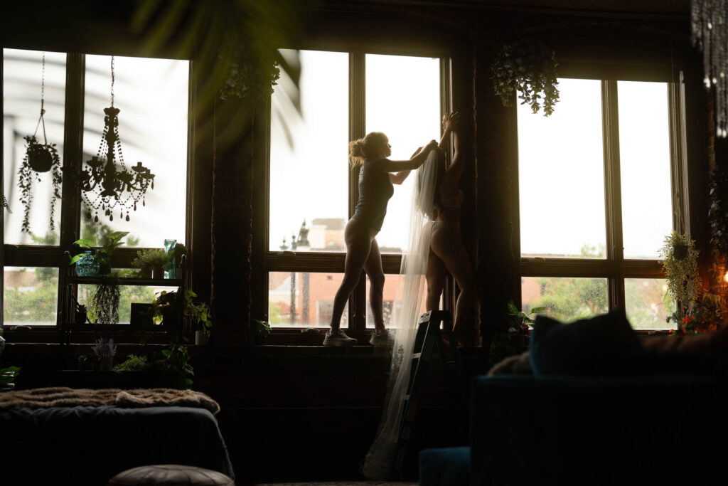 Behind-the-scenes moment of a stylist adjusting a bride’s veil on a step ladder in front of large loft windows at Elevation Boudoir