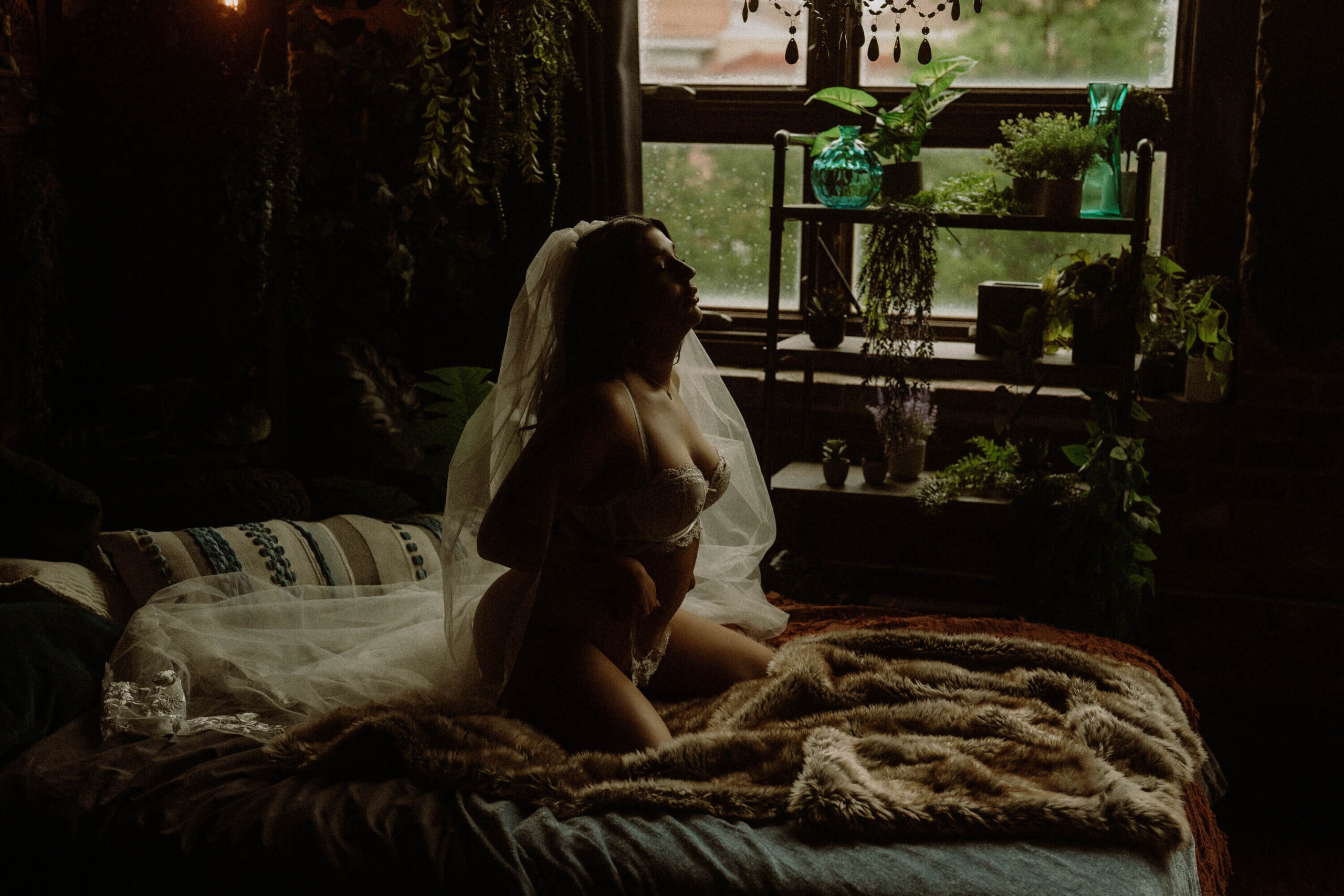 Bride kneeling in lingerie and veil with boho plants and large window behind her during boudoir shoot in Tuscaloosa