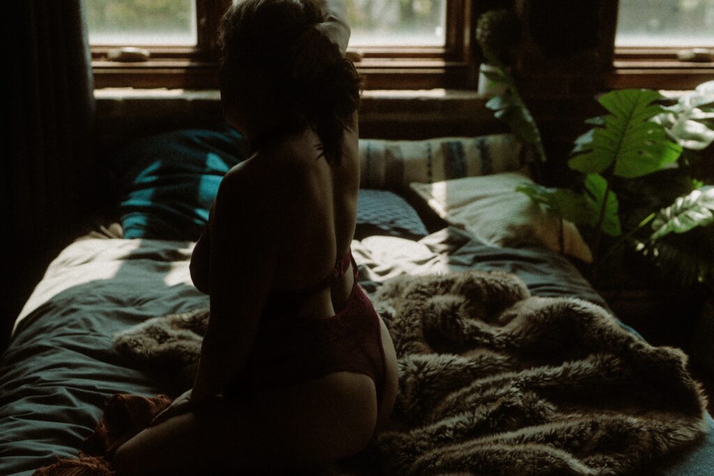 Silhouette of woman sitting on bed in moody light with plants in the background during boudoir session