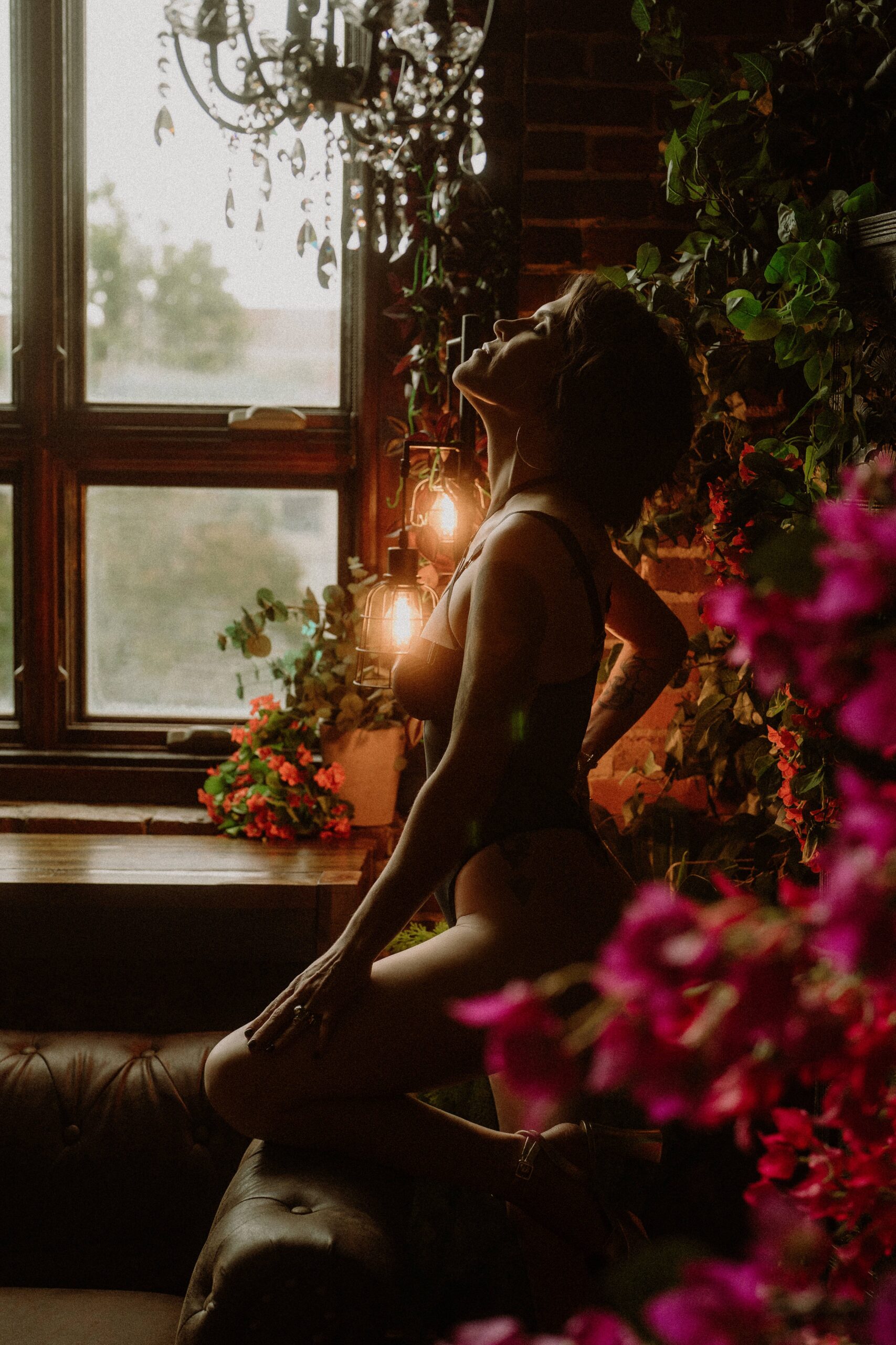 A woman beside a tall window under a crystal chandelier with pink flowers in the foreground during Alabama boudoir photography