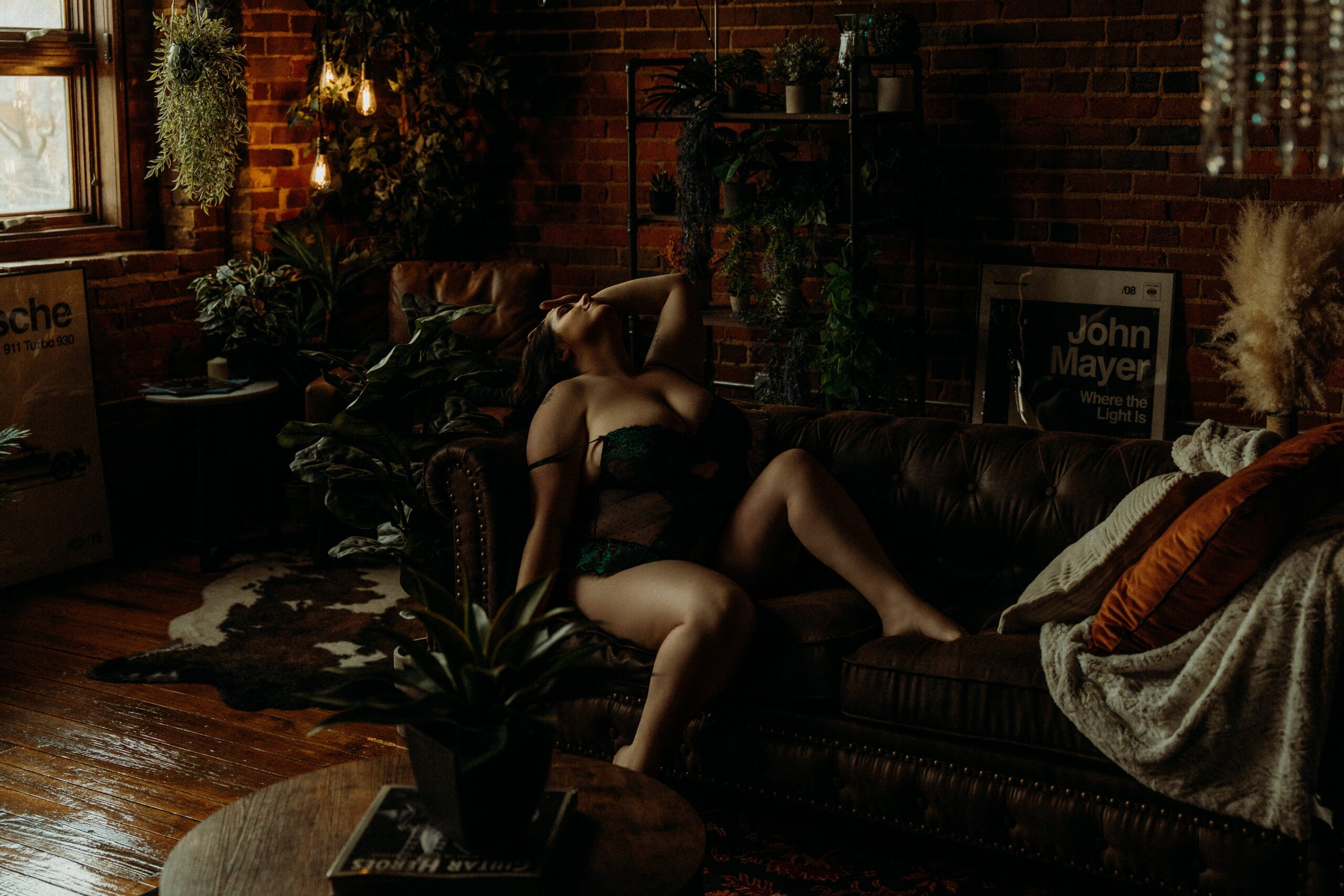 A woman reclining on a dark leather sofa in a warmly lit brick studio with plants and soft Edison bulb lighting during a boudoir session in Tuscaloosa, Alabama