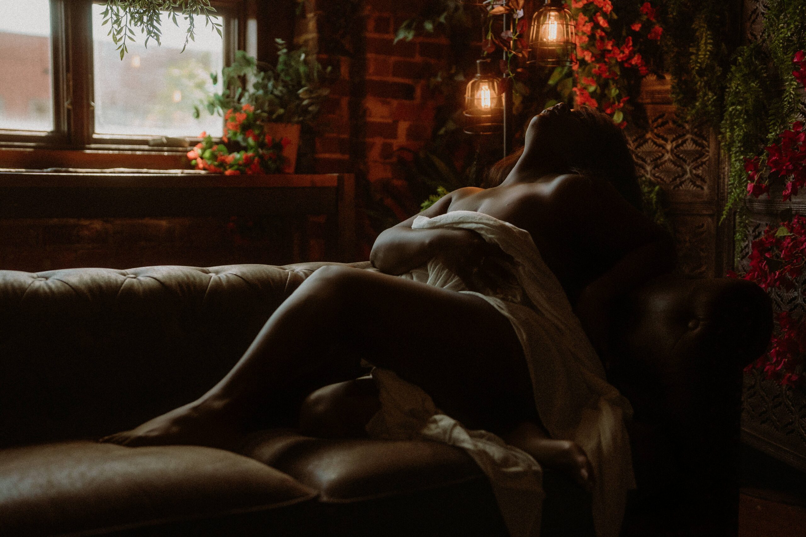 A woman reclining on a leather sofa draped in white fabric beneath warm lanterns and red flowers at a downtown Tuscaloosa boudoir studio