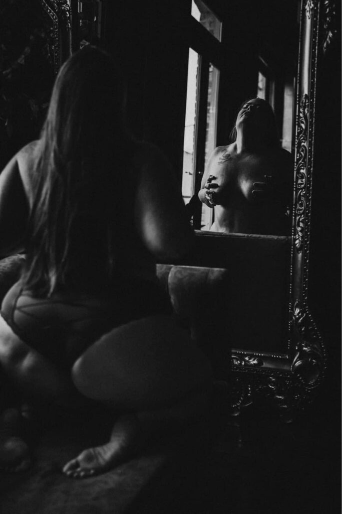 Black and white image of a women holding her breast while posing in front of a mirror in the studio at Elevation Boudoir