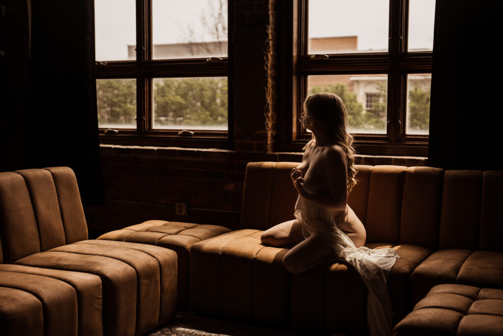 Blonde woman siting on a brown couch holding a white sheet to her chest for boudoir photography in Tuscaloosa AL at Elevation Boudoir Studios