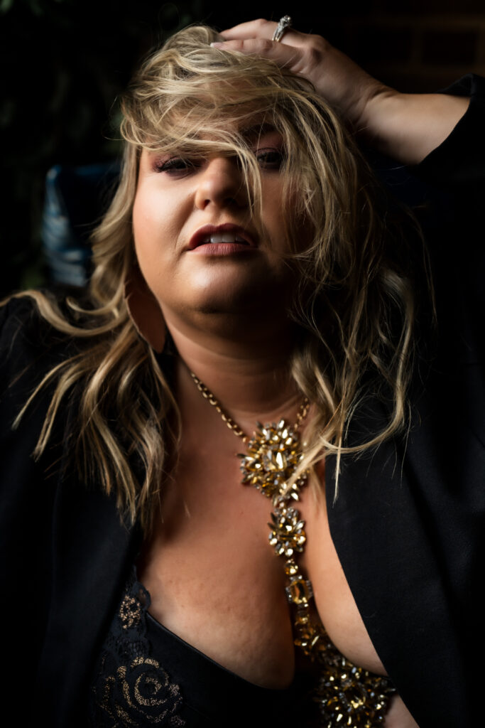 Blonde woman in a yellow/gold body chain and black blazer pushing hair into her face in Elevation Boudoir's Studio